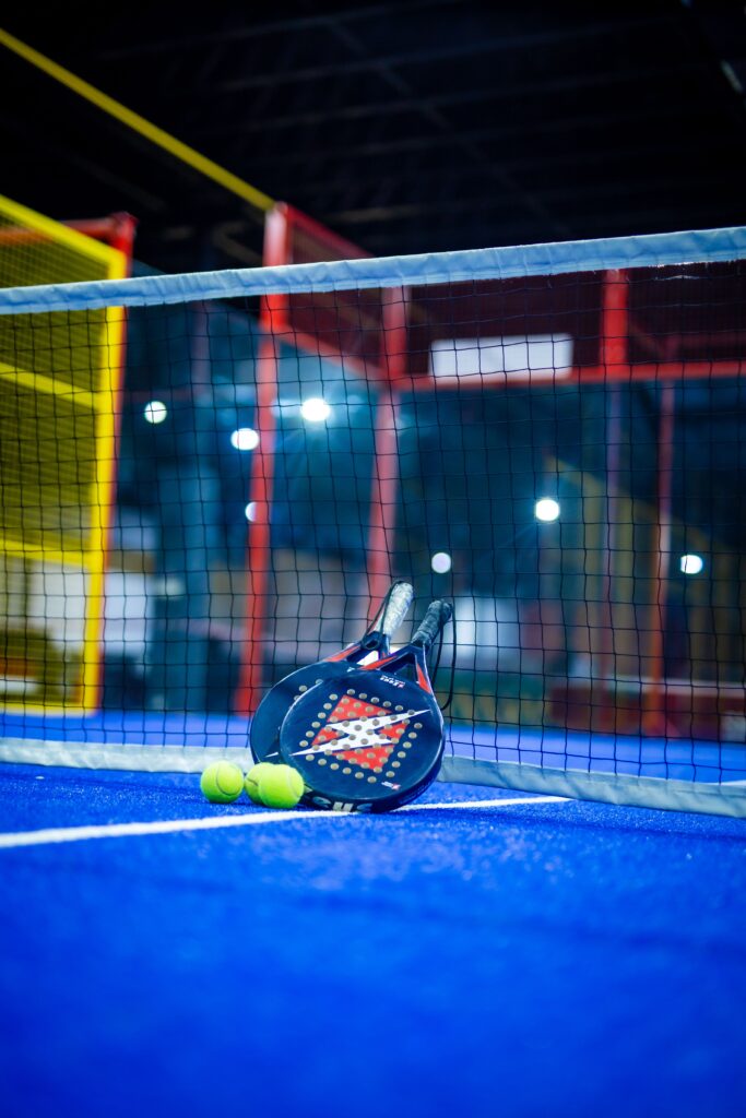 Lapangan padel berwarna biru dengan raket dan bola kuning, yang menonjolkan suasana olahraga yang dinamis di dalam ruangan.